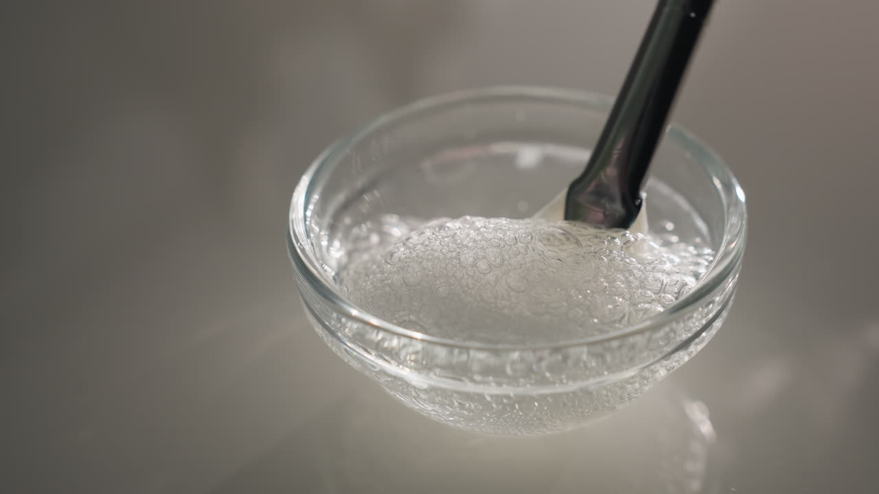 White gel with bubbles lifted by face brush from transparent bowl under soft light to reveal texture and consistency for skincare treatment prep and professional facial application detail