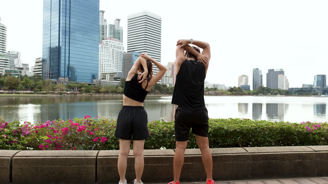 市街のスカイラインを背景に、若い男女が湖畔でオーバーヘッドトライセプスストレッチをしている。