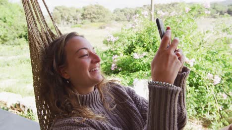 mujer caucásica sonriente acostada en una hamaca usando un teléfono inteligente en un jardín de campo soleado