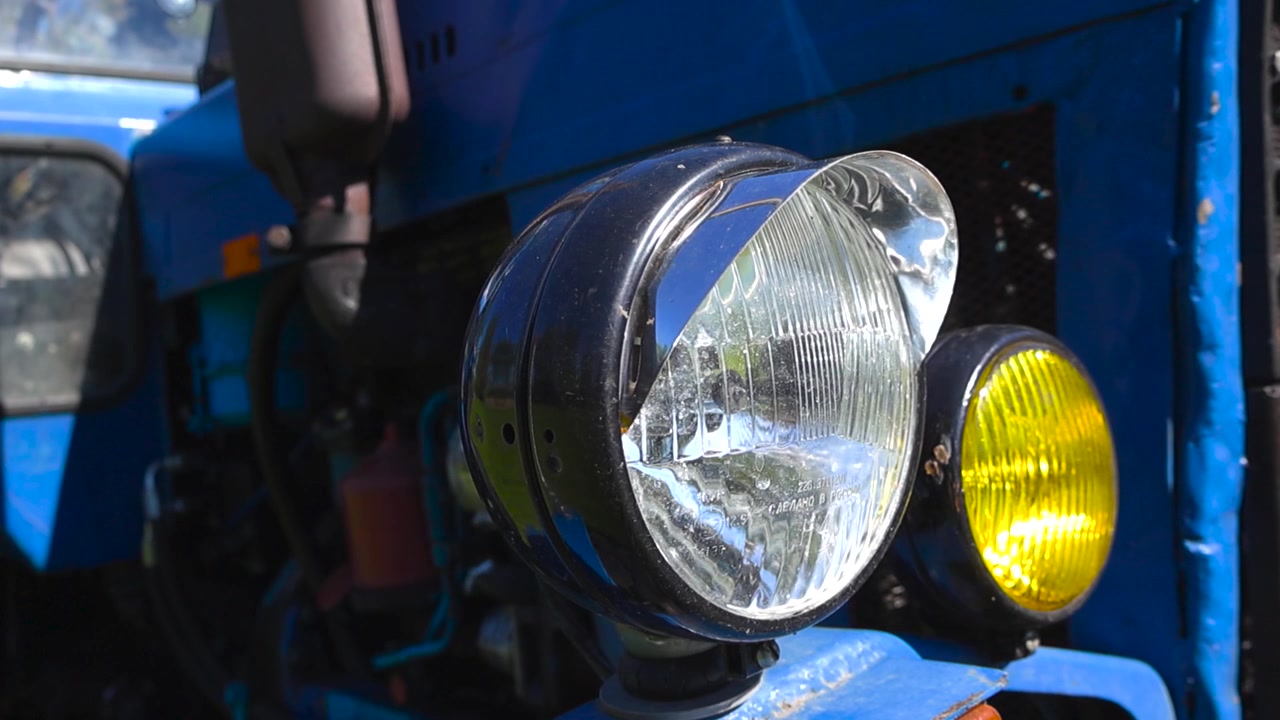 Close up view of a classic blue tractor's chrome headlight and yellow fog lamp, with the camera slowly revealing the details of the old, weathered engine on a sunny day in the countryside