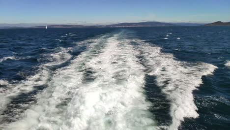 rear trail of the wake of the boat sailing with the land and a sailboat in the background on a sunny day of blue sky in the rías baixas, blocked shot backwards, pontevedra, galicia, spain