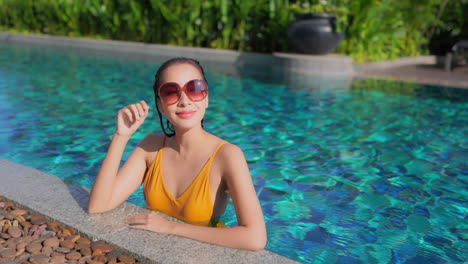 a pretty young woman fresh from a dip in the pool leans along the pool edge and adjust her sunglasses