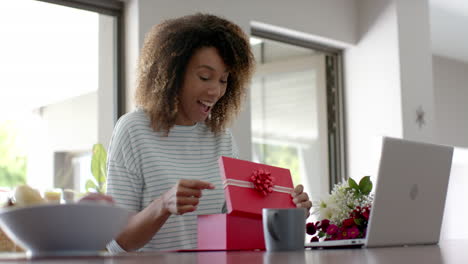 Glückliche-Biracial-Frau-Mit-Geschenk-Und-Blumen,-Die-Zu-Hause-Einen-Videoanruf-Auf-Dem-Laptop-Führt,-Zeitlupe