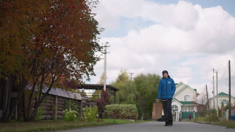 kid in blue jacket strolling thoughtfully along a residential street carrying shopping bag, background features autumn trees, suburban houses, utility poles, and someone walking in the distance