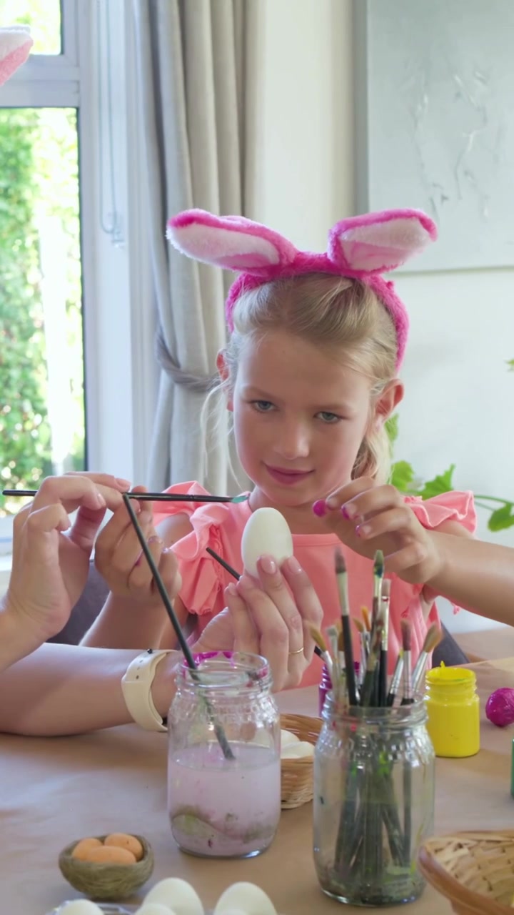 Video verticale: Una donna con un top rosa e orecchie da coniglio, illuminata dal sole, guida una bambina a dipingere uova a casa con i colori.