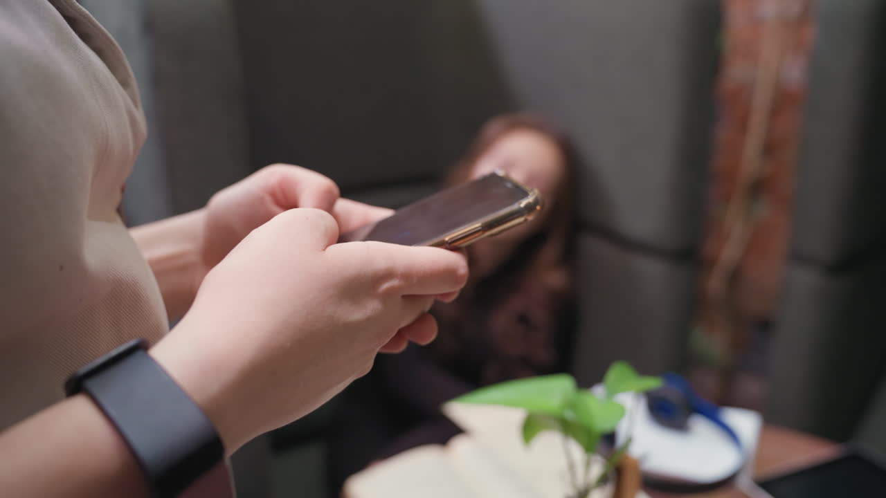 Close-up of woman standing while holding smartphone with both hands and wearing black wristwatch, blurred background shows seated person, table with open book, notepad, and headphones visible