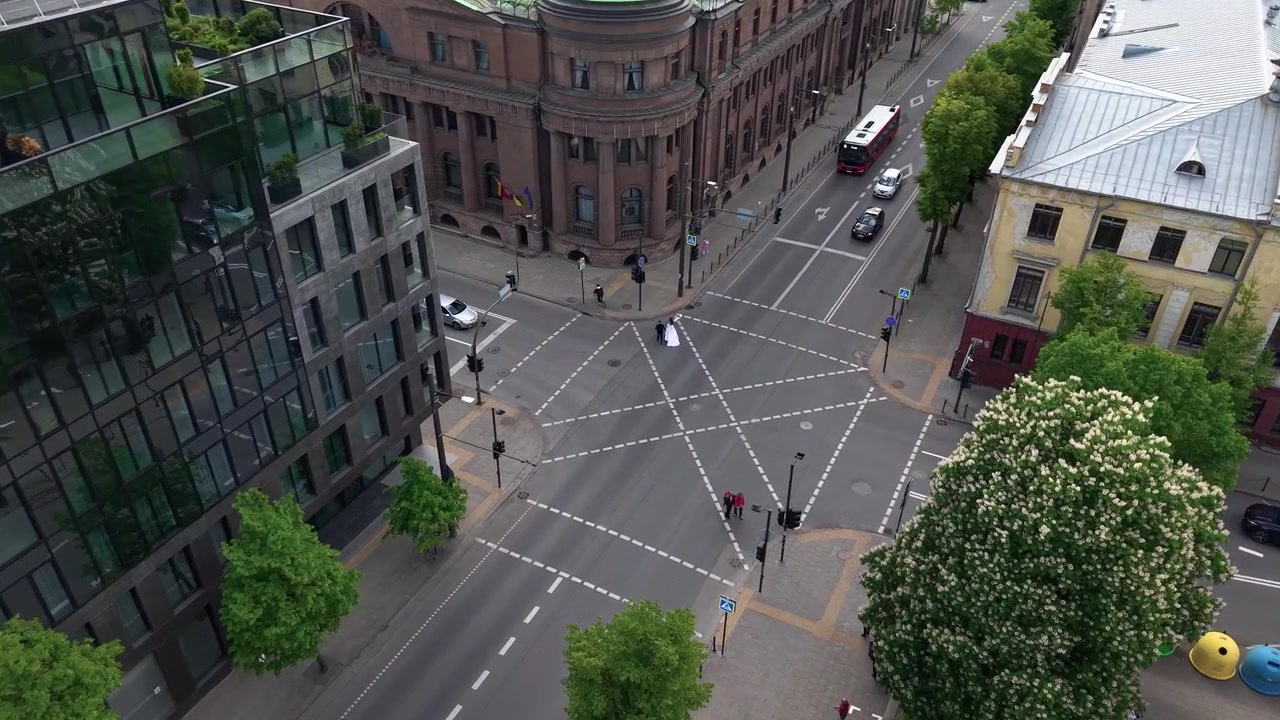 Dronevideo fanger Litauens Bank i Kaunas set ovenfra, med en elegant rundkørsel, fodgængerovergange, moderne glasfacade, trafik, fodgængere, frodige træer og klart dagslys.