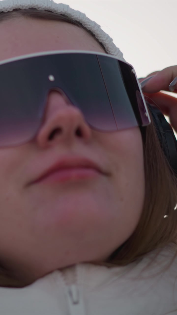 Vista ravvicinata di una donna che indossa grandi occhiali da sole scuri e regola le sue cuffie nere. La donna indossa una giacca bianca con zip e ha lunghi capelli castani, visibili contro uno sfondo di cielo nuvoloso