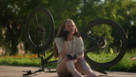 young lady seated next to her upside-down bike, answering phone call with concerned and worried expression, surrounded by lush greenery and vibrant trees in a beautiful outdoor park setting