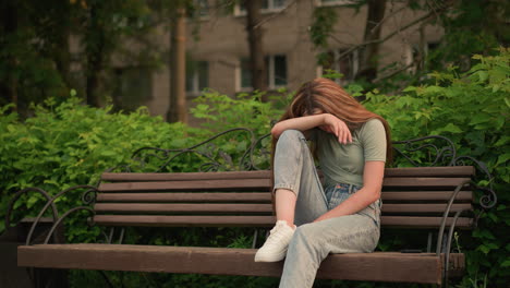 woman in jeans and white sneakers sits on wooden bench in park, resting head on arm with somber, reflective posture, surrounded by lush greenery, trees, expressing solitude