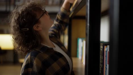 happy girl student with curly hair wearing glasses puts a book on a shelf in the library