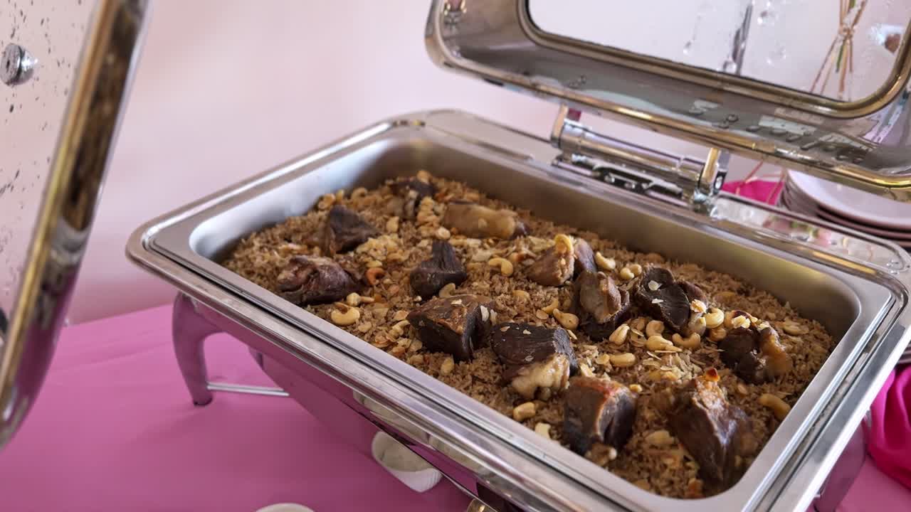 Vista ravvicinata di un tradizionale piatto di agnello e riso guarnito con noci tostate, servito in una scaldavivande in acciaio inossidabile in un allestimento di catering a buffet al coperto con piatti sullo sfondo