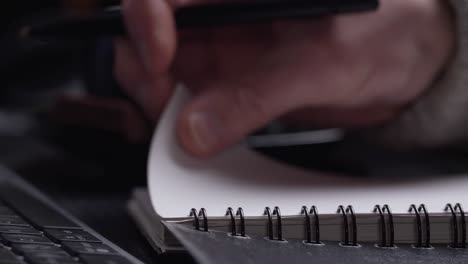 Close-up-of-man's-hands-writing-in-spiral-notepad-placed-on-wooden-black-desktop