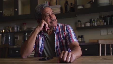 Smiling-senior-mixed-race-man-sitting-in-kitchen-in-thought