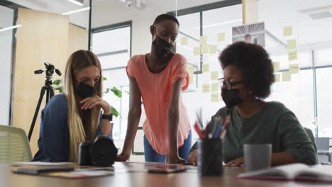 Three-diverse-female-creative-colleagues-wearing-face-masks-in-discussion-at-a-table-in-office