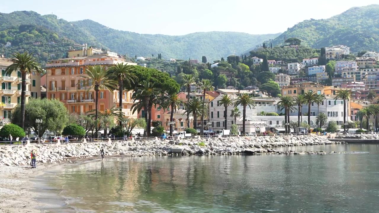 Ampia inquadratura del pittoresco lungomare di Rapallo, della passeggiata fiancheggiata da palme, delle ville color pastello, del tranquillo Mar Ligure e delle colline lussureggianti. Piena luce del giorno, architettura vivace, acqua placida, atmosfera mediterranea serena.