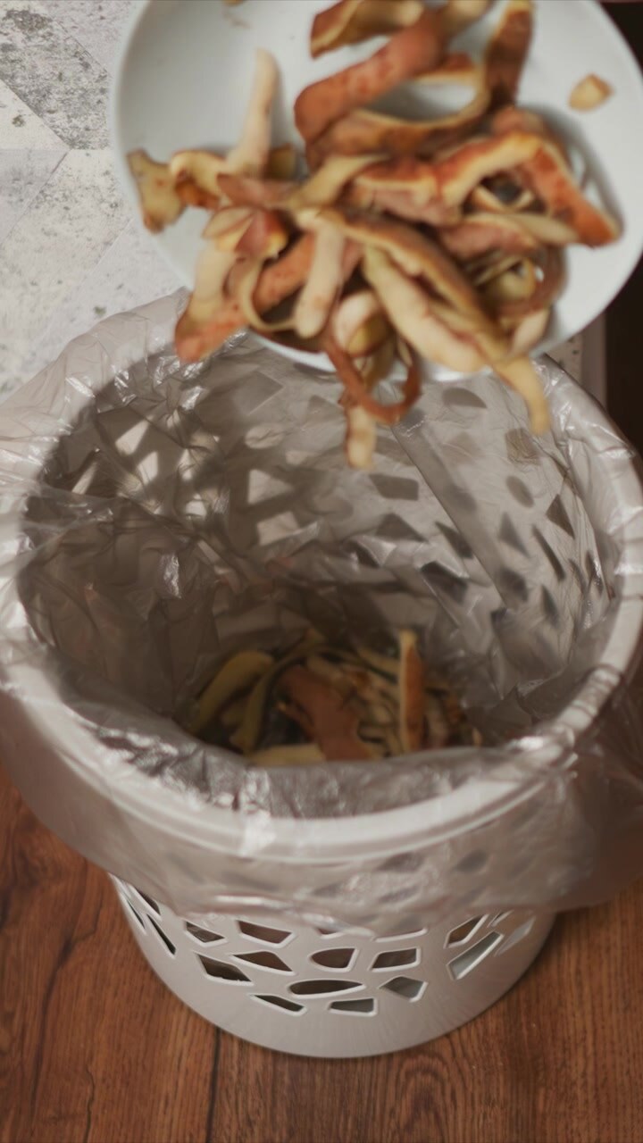 Un bidone della spazzatura bianco con un sacco di plastica si trova su un pavimento di legno accanto a una sedia. Scarti di cibo vengono gettati nella parte superiore aperta del sacco, riempiendolo gradualmente. Smaltimento dei rifiuti.