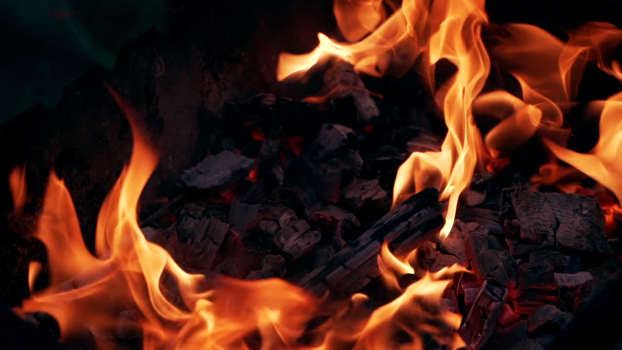 Orange flame and embers of burnt-out fire on dark background. Smoldering logs in small fire outdoors. Glowing fire. Close-up.