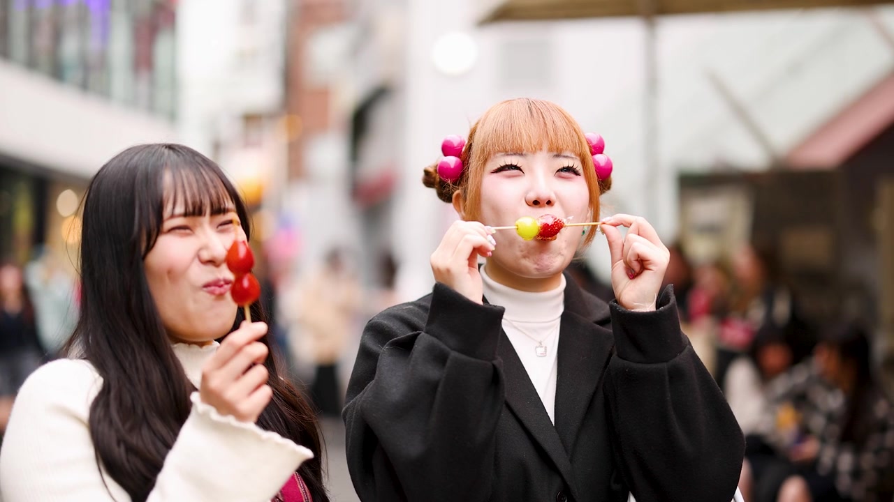 賑やかな東京の露店市場を歩きながら、飴がけフルーツ串を楽しむ若いアジア人女性たち