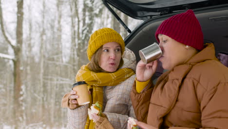 happy couple drinking and eating sandwich while sitting in car boot on a cold winter day