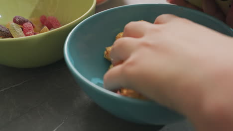 close hand view of person taking flavored popcorn from blue bowl on dark surface with green bowl filled with colorful candy in background, snack time scene with vivid colors and textures