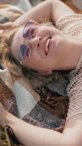 mujer feliz con camiseta y gafas de sol acostada en la sábana al aire libre, sonriendo y tocando la hierba, momento de relajación en la naturaleza con cálida luz solar y atmósfera pacífica