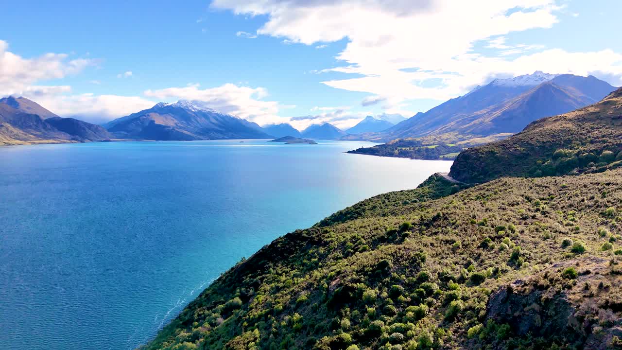 Drone glides above turquoise lake, lush hills, and distant mountains under bright daylight skies