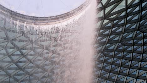 a dramatic indoor waterfall pours from a circular skylight, framed by geometric glass panels and natural daylight, captured in a smooth upward camera pan