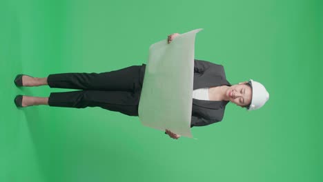 full body of smiling asian female engineer with safety helmet looking at blueprint in the green screen studio