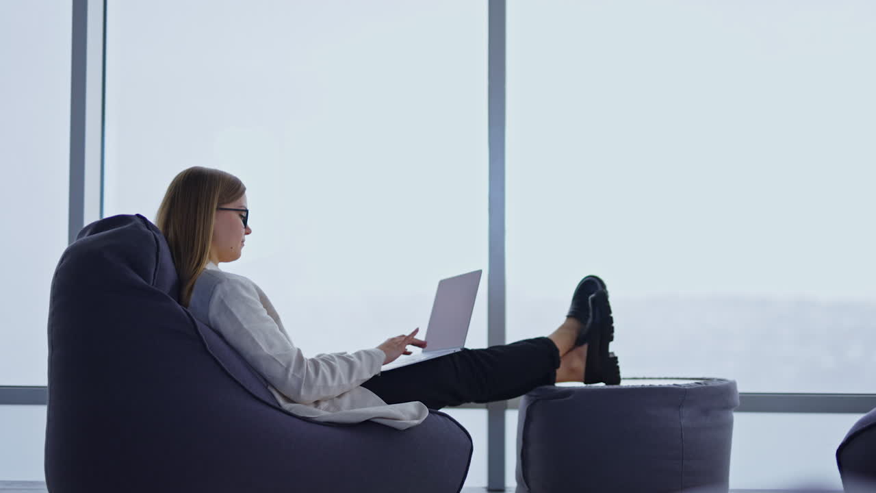 Comfortable working atmosphere in a bean bag chair. Lady typing on the laptop placed on her laps. White window backdrop. Side view.