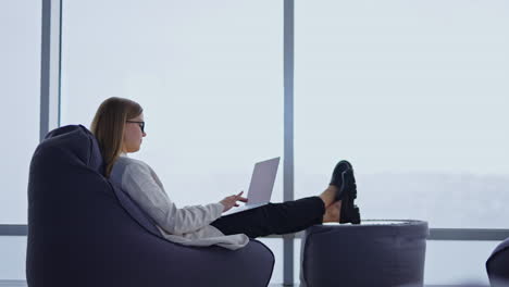 comfortable working atmosphere in a bean bag chair. lady typing on the laptop placed on her laps. white window backdrop. side view.