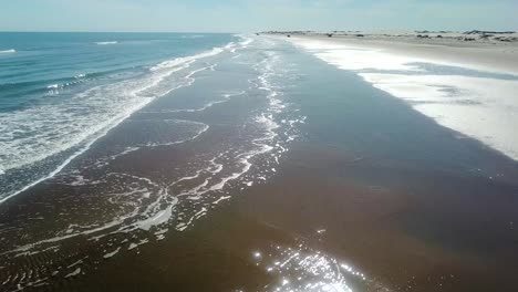 Aerial-drone-view-following-the-shoreline-and-surf-at-low-tide-on-a-gulf-coast-barrier-island-on-a-sunny-afternoon,-fisherman-and-vehicles-in-the-distance---South-Padre-Island,-Texas