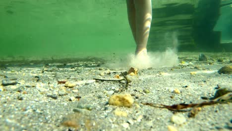 Underwater-clip-of-childs-feet-walking-on-beach-bottom-in-beautiful-sunlight---Sun-giving-glimmering-light-at-seabed---Static-underwater-with-camera-laying-on-seabed