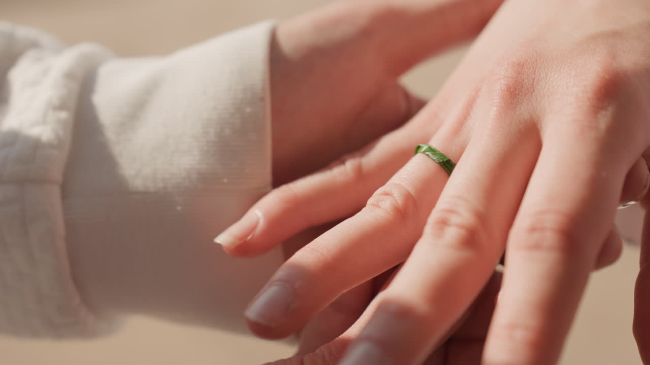 Dito sabbioso con anello verde su mano caucasica. Primo piano sulla spiaggia mostra sabbia texturizzata e fascia di smeraldo che scivola sull'anulare, bagliore illuminato dal sole e movimento delicato catturano un gioioso momento di matrimonio.
