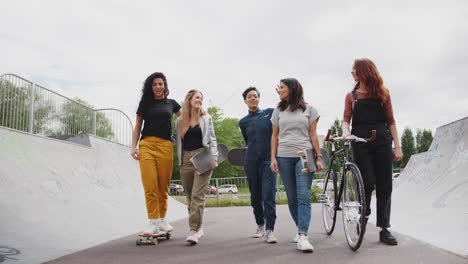 female friends with skateboards and bike walking through urban skate park