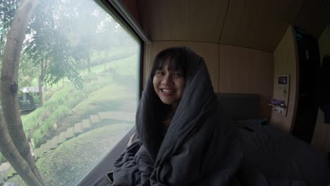 Indonesian girl in a cabin wrapped in a blanket, smiling and relaxing with nature views