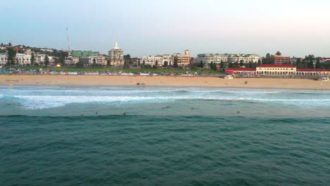 Drone-tracking-left-along-Bondi-beach-with-surfers-in-the-water-and-Lifeguards-on-the-beach,-Sydney-Australia