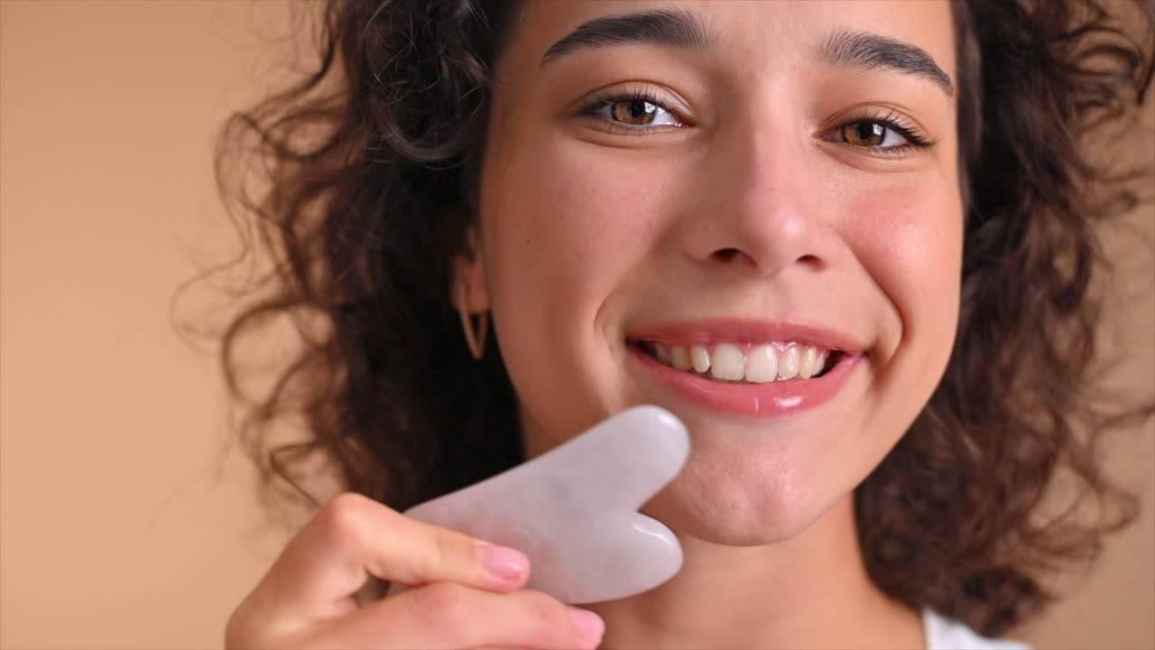 A young caucasian smiling woman is doing a facial massage using Gua Sha, looking into the camera. Slow motion