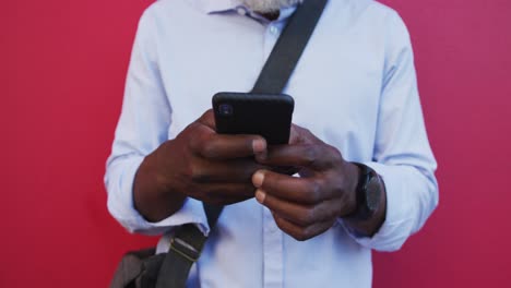 Portrait-of-african-american-senior-man-using-smartphone-while-standing-against-pink-wall