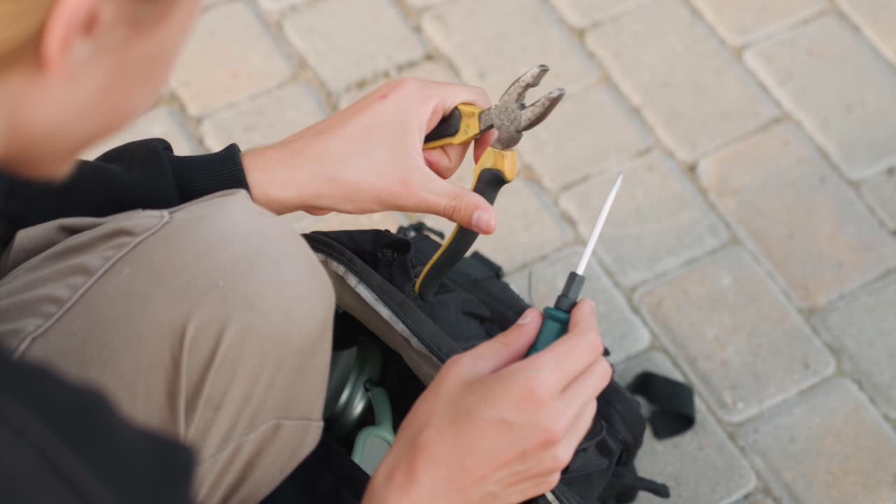 Una donna caucasica bionda sistema gli attrezzi nello zaino sulla pavimentazione, seleziona pinze e cacciavite, organizza i componenti prima di un progetto, preparazione paziente e mani ferme, primo piano dall'alto dell'attrezzatura.