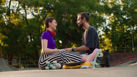 Side-shot-of-a-brunette-guy-in-a-gray-t-shirt-and-a-short-haired-girl-in-a-purple-top-and-striped-pants-are-sitting-on-the-floor-in-a-skate-park-and-chatting.-The-guy-touches-the-cheek-of-the-girl