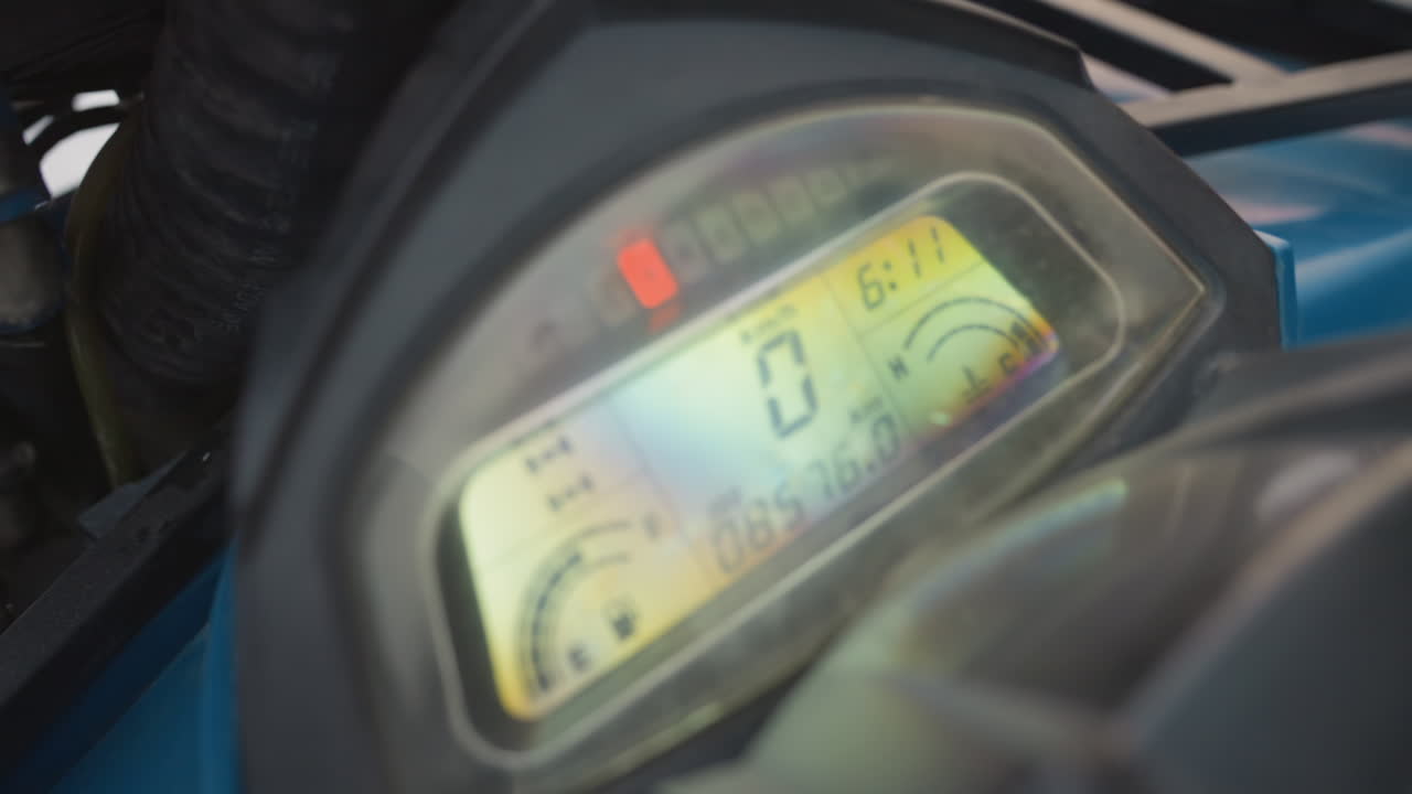 Atv dashboard closeup showing digital display, gloved hand adjusts controls during preride inspection on blue quad, speedometer reads zero, illuminated indicators and odometer visible, plastic