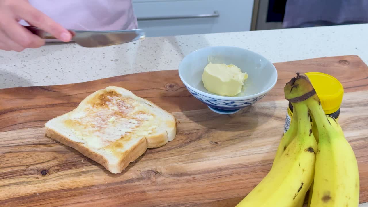 A person spreads butter onto slices of toasted bread on a wooden countertop, with bananas and a butter dish nearby under bright, even lighting