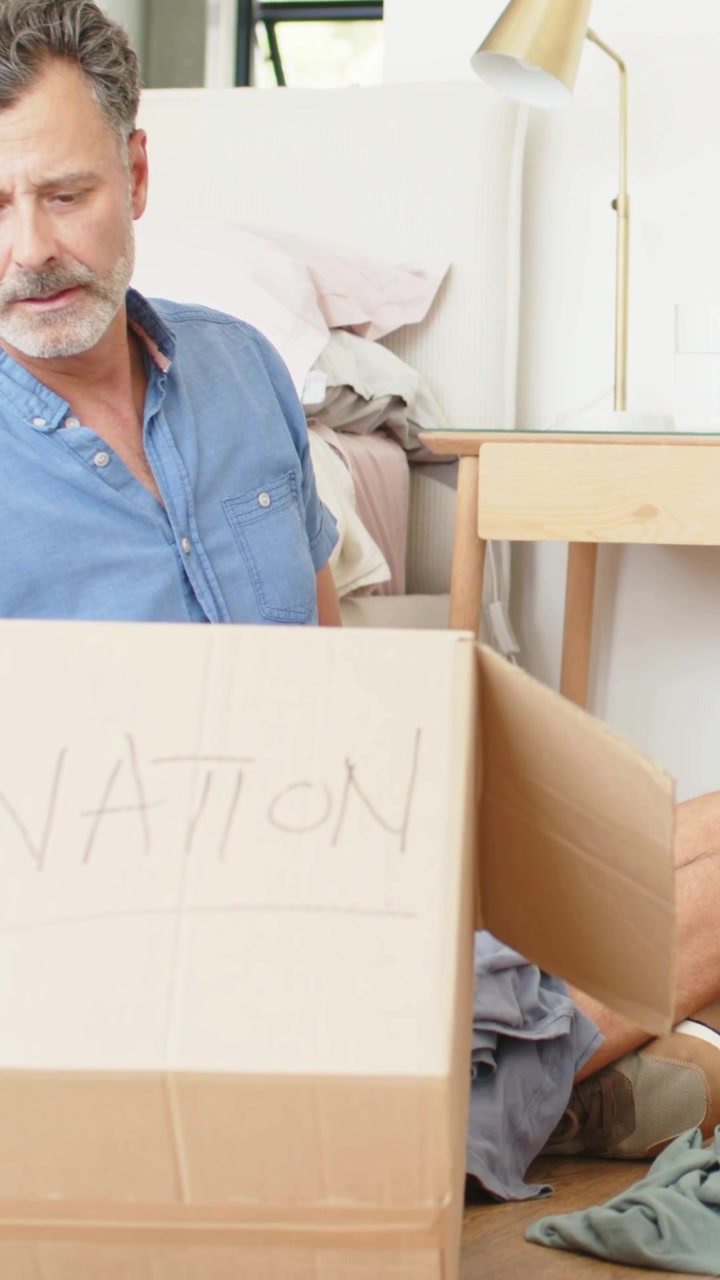 Vertical video: Opening marked box, man wearing blue shirt sorting clothes beside bed for donation