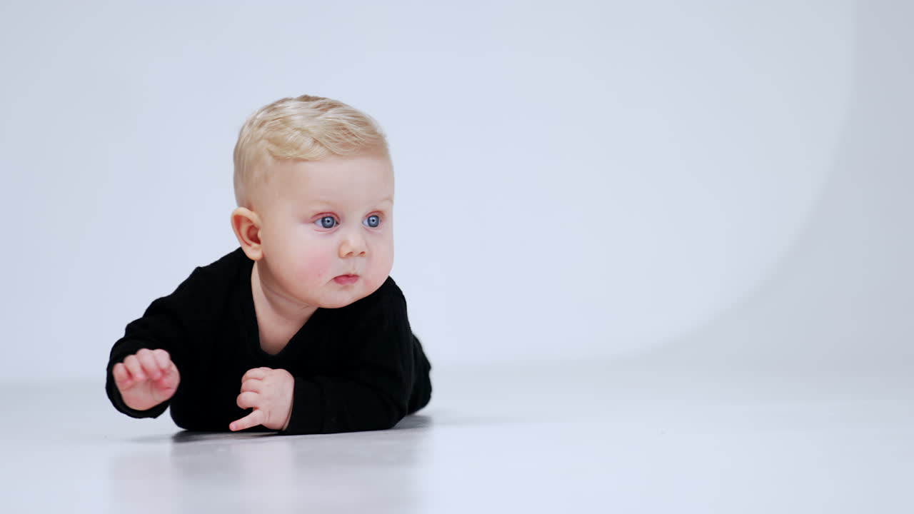 Adorable blond baby boy in black suit lies on the floor. Persistent kid crawls up to camera. White backdrop.