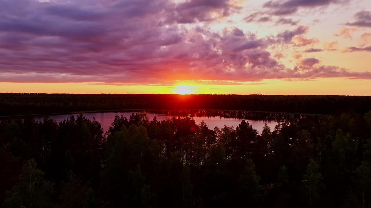 Hehkuva auringonlasku maalaa pilviä tyynen suojärven yläpuolella, tumman metsän ympäröimänä.