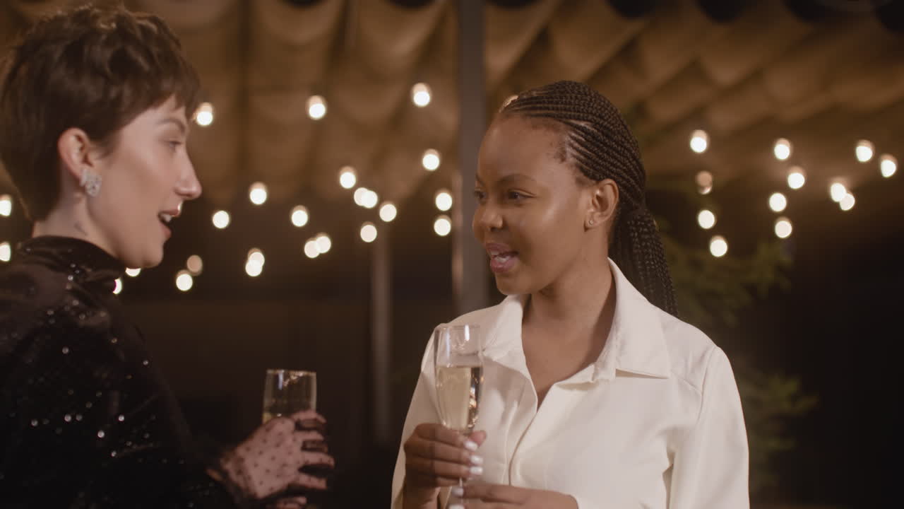 Portrait Of Two Elegant Multiethnic Women With Champagne Glass Talking To Each Other And Then Welcoming Two Men To New Year's Eve Party