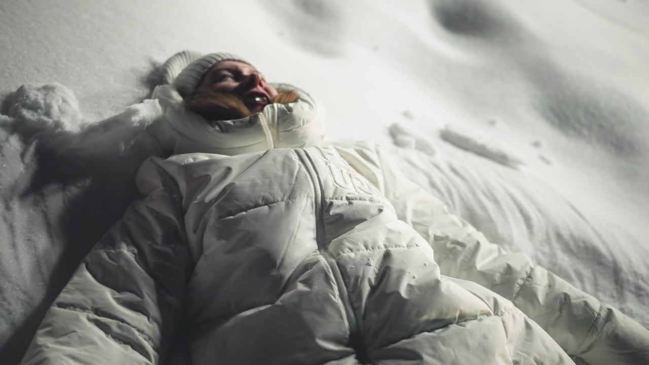 Student lying on snowy surface, smiling and playing joyfully under night sky in winter attire, arms stretched out and body surrounded by fresh snow, capturing carefree moment of youthful enjoyment