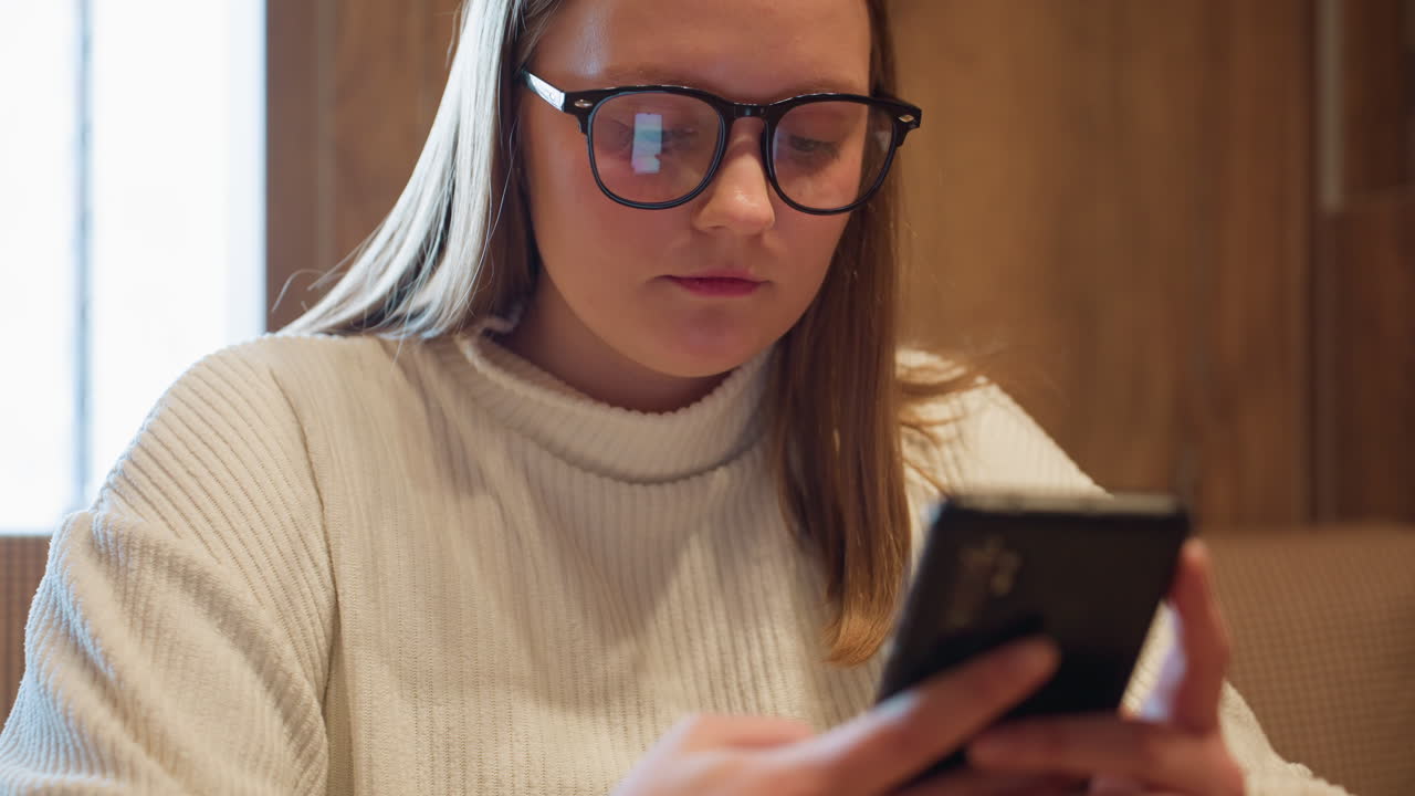 Close up of girl in white sweater and glasses focused on smartphone while chatting, phone screen clearly reflecting in lenses, creating warm indoor ambiance with wooden background