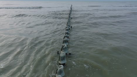 Aerial-birdseye-view-of-an-old-wooden-pier-at-the-Baltic-sea-coastline,-overcast-winter-day,-white-sand-beach-covered-in-snow,-ice-on-wood-poles,-calm-seashore,-wide-drone-shot-moving-back-low